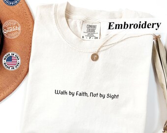 May include: Cream-colored t-shirt with the embroidered text "Walk by Faith, Not by Sight." The shirt features a crew neck and short sleeves. A gold-colored necklace with a pendant is draped on the shirt. The word "Embroidery" is printed on the shirt.