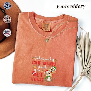 Peut inclure: Un t-shirt corail à col rond avec la phrase brodée "Jolliest punck of Chip Munks this side of the Nut House" et un motif de dessin animé d'écureuil. Un collier en or avec un pendentif est drapé sur le t-shirt.
