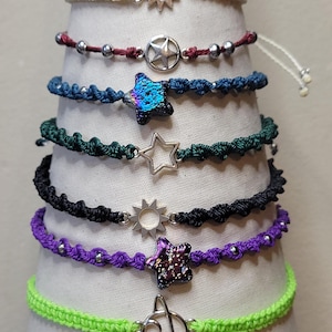 May include: A collection of handmade macrame bracelets in various colors, including cream, burgundy, blue, green, black, purple, and neon green. Each bracelet features a unique charm, such as stars, suns, and a planet design, and is adorned with silver beads.