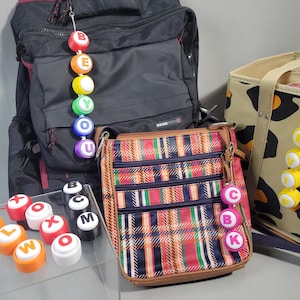Pode incluir: Uma coleção de bolsas e acessórios, incluindo uma mochila preta, uma bolsa transversal xadrez e uma sacola com estampa de leopardo. Pingentes de letras coloridas soletram palavras, presas às bolsas. Outros pingentes de letras são exibidos em uma superfície transparente.