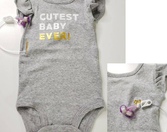 May include: A gray baby bodysuit with ruffled sleeves and the text "Cutest Baby Ever!" in white and gold lettering. The bodysuit has a snap closure at the bottom and a medical device attached.