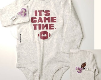 May include: Light gray baby bodysuit with long sleeves. The bodysuit features the text "IT'S GAME TIME." in maroon, along with a maroon football graphic. A medical device is attached to the sleeve. The bodysuit is from the brand Carter's.
