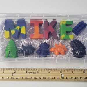 May include: A clear plastic container with the name "MIKE" spelled out in colorful crayons. The crayons are shaped like letters and various objects, including a Lego brick and dinosaurs. The container is filled with white paper shreds.