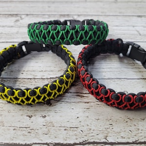 May include: Three paracord bracelets in black with accent colors of green, yellow, and red. Each bracelet features a woven design and a black plastic buckle clasp. The bracelets are displayed on a light wood-grain surface.