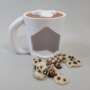 Puede incluir: Una taza blanca con asa, llena de líquido marrón y cubierta con malvaviscos blancos. Varias galletas con forma de vaca con manchas marrones están esparcidas alrededor de la taza. La taza tiene una sección recortada.