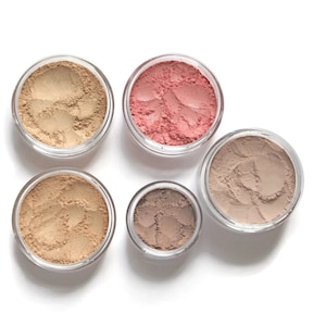 May include: Five small round containers of pressed powder makeup. The containers are clear plastic and the powders are various shades of brown, beige, and pink.