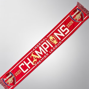 以下が含まれることがあります： アーセナルのロゴと「CHAMPIONS」と「PREMIER LEAGUE」の文字が入った赤いスカーフ。スカーフには2025年と2026年が表示されています。スカーフの両端にはフリンジがあり、アーセナルのロゴが両端にあります。