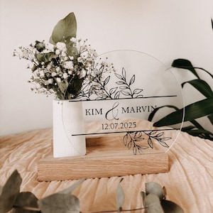 Könnte beinhalten: Runder Hochzeitsanzeiger aus transparentem Acryl mit schwarzem Blumendesign. Er trägt die Namen "KIM & MARVIN" und das Datum "12.07.2025". Das Schild steht auf einem Holzsockel, mit einer weißen Blumenvase.