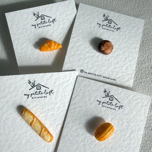 May include: Four miniature food items on white cards. A croissant, a round pastry, a baguette, and an oblong pastry are displayed. Each card has the text "my petite loft miniatures" and the Instagram handle "@my.petite.loft.miniatures".