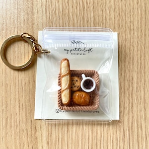 May include: A keychain featuring a miniature scene of a baguette, pastry, and a cup of coffee in a small wicker basket. The keychain is packaged in a clear plastic bag with the text "my petite loft miniatures" printed on the bag.