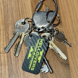 May include: A set of keys on a wooden surface, held together by a silver carabiner and key rings. A black rectangular keychain with the word "HELLDIVERS" in yellow is also attached to the key ring.