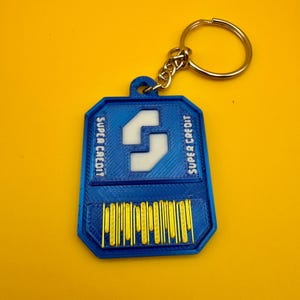 May include: A blue and yellow keychain with a white "S" logo and the text "SUPER CREDIT" on both sides. The keychain has a gold metal ring.