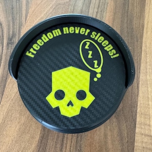 May include: A black coaster with a carbon fibre texture and a raised edge. The coaster features a neon yellow skull design and the phrase "Freedom never sleeps!" in a circular pattern. A thought bubble with "Zzz" is also present.