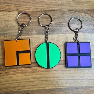 May include: Three keychains featuring different geometric shapes. The first keychain has a square shape with an orange background and a black outline. The second keychain has a circle shape with a green background and a black line down the middle. The third keychain has a square shape with a purple background and a black outline.
