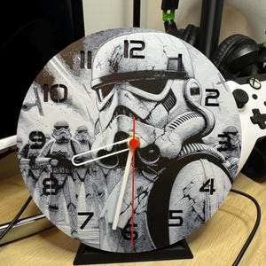 May include: A round, black and white clock with a Star Wars Stormtrooper design. The clock has a black stand and white hands. The numbers on the clock face are black.