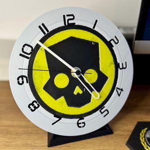 May include: A circular clock with a white face, black numerals, and hands. The clock's face displays a black skull design on a yellow backdrop. It's mounted on a black stand, resting on a wooden surface.