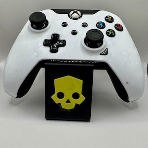 May include: White Xbox controller with black buttons and a skull logo on the stand. The skull is yellow with black eyes.