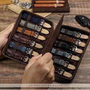 Puede incluir: Un estuche de viaje de cuero marrón para correas de reloj, abierto para mostrar varias correas de reloj en tonos azules, marrones y grises. El estuche tiene cremallera y está forrado con un material marrón suave. El texto "GEMHUNTERCRAFTS" es visible en la parte inferior.