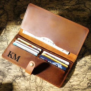 May include: A brown leather long wallet featuring a snap button closure, multiple card slots, and a large bill compartment. The exterior is engraved with the letters KM. The wallet is open, revealing several credit cards and paper currency inside.