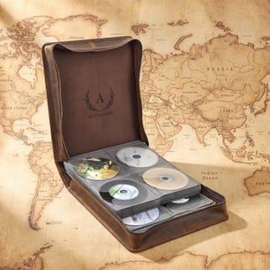 May include: A brown leather CD storage case with a zipper closure, featuring an open interior with multiple clear plastic sleeves holding several compact discs. The inside flap is embossed with a laurel wreath, the letter A, and the name ALEXANDER.