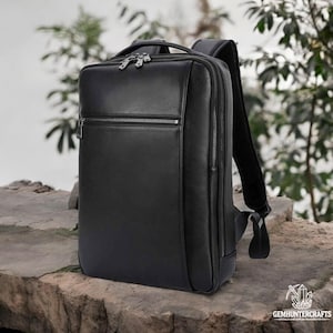 Könnte beinhalten: Ein schwarzer Lederrucksack mit einer Reißverschlusstasche vorne und verstellbaren Trägern. Der Rucksack steht auf einer steinernen Oberfläche.
