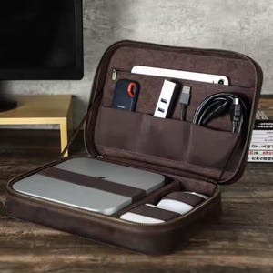 May include: A brown leather carrying case with a zip closure. The case is open and has several compartments for electronics and accessories. The case is sitting on a wooden surface.
