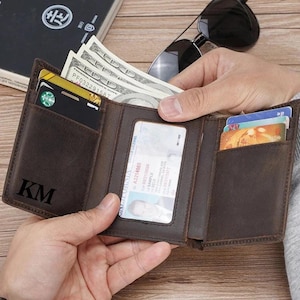 May include: A dark brown leather tri-fold wallet featuring the embossed initials KM on the front left panel. The wallet is open, revealing multiple slots containing US dollar bills, a driver's licence in a clear window, and various credit cards.