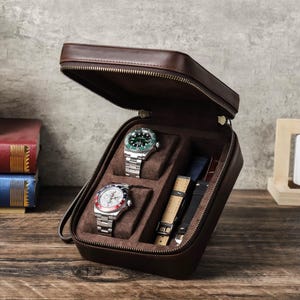 Könnte beinhalten: Eine braune Leder-Uhrenbox mit Reißverschluss, geöffnet, um zwei Uhren und zwei Armbänder zu zeigen. Eine Uhr hat eine grüne Lünette und ein grünes Zifferblatt, die andere ein weißes Zifferblatt mit roten und blauen Akzenten. Das Etui ist mit braunem Filz ausgekleidet.