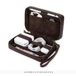 May include: Open brown leather tech organizer with a zipper closure. Inside are white charging accessories, a stylus, and a smartwatch. The organizer has a wrist strap and is on a white background.