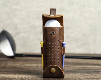 May include: A brown leather golf ball holder with a white golf ball inside. The holder has a strap and a perforated design. There are two yellow and two purple golf tees sticking out of the holder.