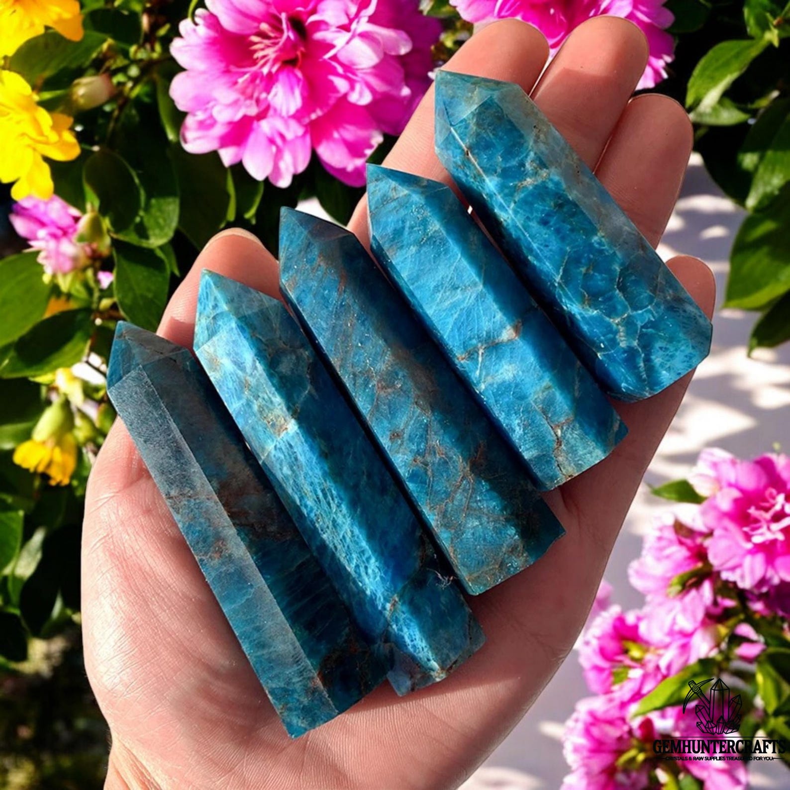 Polished blue Apatite towers held in a hand, used to enhance Gemini traits, with bright pink and yellow flowers in the background.