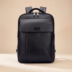 May include: A black textured leather backpack featuring a front flap pocket with the letters KM embossed in the center. The bag includes a vertical zippered compartment on the front, a top handle, and adjustable shoulder straps with silver hardware.