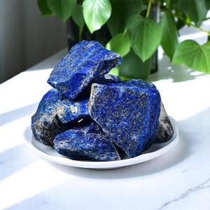 Puede incluir: Una colección de piedras de lapislázuli azul rugosas con motas doradas. Las piedras están dispuestas en una pila sobre un plato blanco.