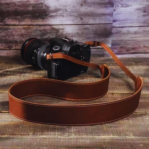 Puede incluir: Una correa de cámara de cuero marrón con un borde cosido. La correa está envuelta alrededor de una cámara negra con una lente grande.
