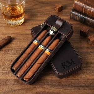 Könnte beinhalten: Ein dunkelbraunes Zigarrenetui aus Leder mit Metallverschluss und den eingeprägten Buchstaben KM. Das Etui enthält drei braune Zigarren mit orange-schwarzen Cohiba-Banderolen. Das Etui ist geöffnet und zeigt ein weiches Innenfutter.