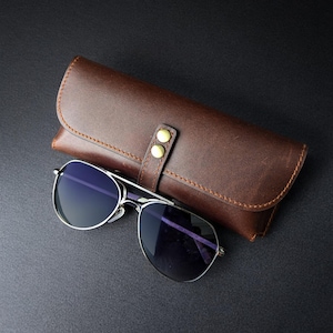 Könnte beinhalten: Aviator-Sonnenbrille mit silbernem Rahmen und dunklen Gläsern neben einem braunen Lederetui. Das Etui hat einen Klappverschluss, der mit einem Riemen mit zwei goldfarbenen Knöpfen befestigt ist. Die Sonnenbrille und das Etui liegen auf einer dunklen Oberfläche.