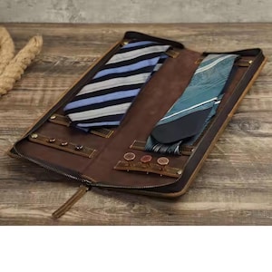 May include: A brown leather tie case with a zipper closure. The case has two tie slots and a section for tie clips. The case is open and two ties are visible, one blue and white striped and one teal.