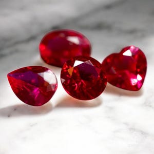 以下が含まれることがあります： 白い大理石の表面に、異なる形と大きさの赤いルビーの宝石が4つあります。