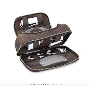 Op de afbeelding: Open bruin lederen tech-organizer met meerdere compartimenten. De organizer is gevuld met witte oplaadaccessoires en een computermuis. De rits is goudkleurig. De tekst "GEMHUNTERCRAFTS" staat onderaan.
