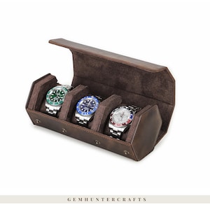 Peut inclure: Un étui à montres en cuir marron avec trois compartiments, chacun contenant une montre-bracelet argentée avec un cadran de couleur différente : vert, bleu et rouge/blanc. L'étui est ouvert, révélant un intérieur doux. La marque "GEMHUNTERCRAFTS" est en bas.