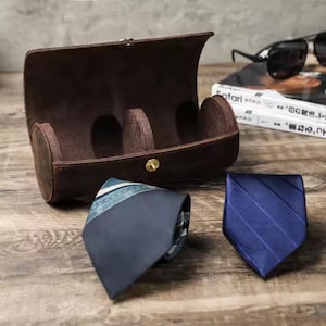 Può includere: Un astuccio cilindrico in pelle marrone con tre scomparti per riporre orologi o altri piccoli oggetti. Due cravatte, una blu e una con un motivo blu e verde, sono appoggiate su una superficie di legno.