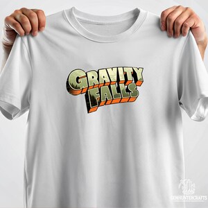 Może przedstawiać: Biała koszulka z grafiką w stylu retro z napisem "Gravity Falls" w kolorze pomarańczowym i zielonym.