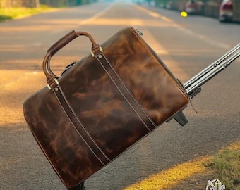 Op de afbeelding: Een bruine leren rolreistas met een bovenste handvat en een intrekbaar handvat. De tas staat op een geplaveide weg met een wazige achtergrond.