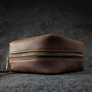 Puede incluir: Un neceser de viaje de cuero marrón con cremallera de latón. El estuche rectangular tiene un diseño cosido y un pequeño lazo en el lateral. El cuero tiene un aspecto envejecido.