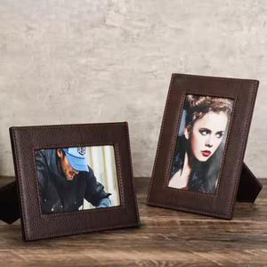 May include: Two brown leather picture frames with stitched edges. The frame on the left has a photo of a person wearing a blue baseball cap. The frame on the right has a photo of a person with long brown hair.