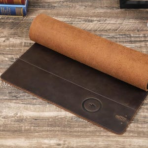 Puede incluir: Una alfombrilla de escritorio de cuero marrón con una almohadilla de carga inalámbrica integrada. La almohadilla de carga tiene un símbolo de rayo. La alfombrilla está enrollada y el cuero es visible.