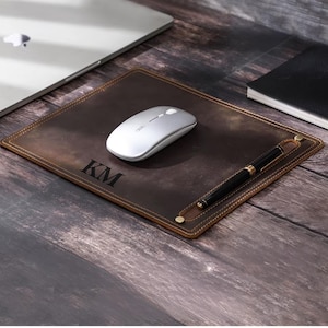 May include: A brown leather desk mat with a pen holder and the initials "KM" embossed on the surface. A silver wireless mouse is on the mat.