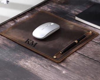 May include: A brown leather desk mat with a pen holder and the initials "KM" embossed on the surface. A silver wireless mouse is on the mat.