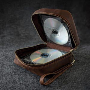 May include: A brown leather CD case with a zipper closure, open to reveal several CDs. The case has a wrist strap and a dark background. The CDs are visible through clear plastic sleeves.