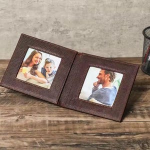Puede incluir: Un marco de fotos de cuero marrón con dos aberturas para fotos. El marco está cerrado y las fotos muestran una mujer y un niño en una abertura y un hombre en la otra abertura.